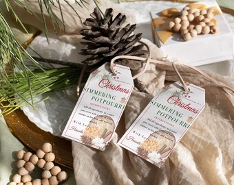 Peut inclure: Deux étiquettes cadeaux blanches sur le thème de Noël. Les étiquettes présentent un design rouge et vert avec une illustration aquarelle d'oranges, de bâtonnets de cannelle et de pommes de pin. Le texte sur les étiquettes indique "Noël" et "Pot-pourri mijoté" avec des instructions d'utilisation. Les étiquettes sont attachées avec de la ficelle et sont posées sur une surface en tissu blanc.