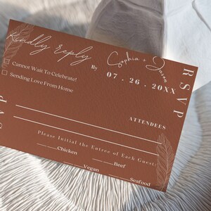 May include: A brown RSVP card with white text. The card has a feather design and the text "Kindly reply" at the top. The text "Sophia & Jason" is written in cursive. The date "07.26.20XX" is written below. The card has a checkbox for "Cannot Wait To Celebrate!" and "Sending Love From Home". The card also has a section for "Attendees" and a list of entree options: "Chicken", "Beef", "Vegan", and "Seafood".