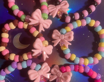 May include: Six colorful beaded bracelets with pink bows. Each bracelet is made with different colored beads and has a pink bow charm attached.