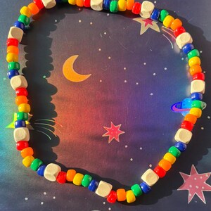 May include: A colorful beaded necklace with a rainbow of colors, including red, orange, yellow, green, and blue. The necklace also features small, square, wooden beads. The necklace is displayed on a space-themed background with stars, planets, and a crescent moon.