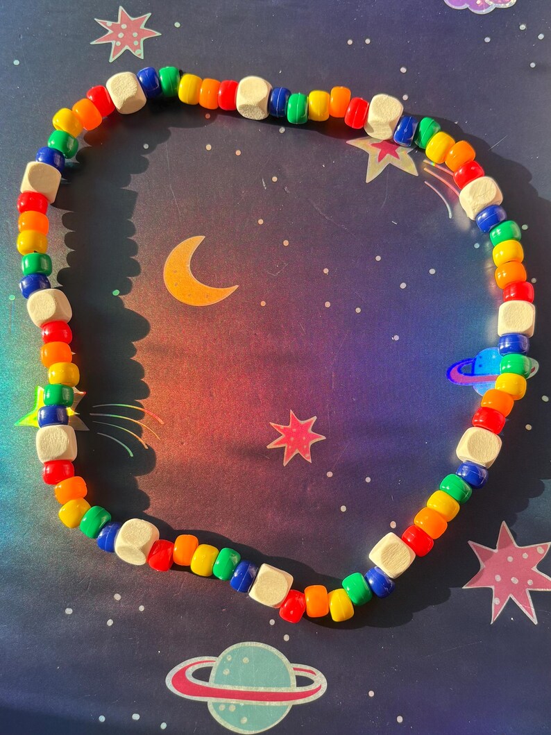 May include: A colorful beaded necklace with a rainbow of colors, including red, orange, yellow, green, and blue. The necklace also features small, square, wooden beads. The necklace is displayed on a space-themed background with stars, planets, and a crescent moon.