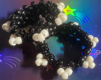 May include: A black and white beaded bracelet. The bracelet is made of small, round beads in alternating colors. The beads are strung together to form a flexible, circular shape. The bracelet is on a colorful background with stars and planets.