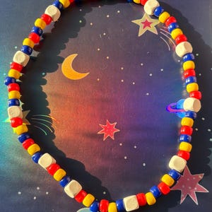 May include: A colorful beaded necklace with red, yellow, blue, and white beads. The necklace is arranged in a circular shape, with a space-themed background featuring stars, planets, and a crescent moon.