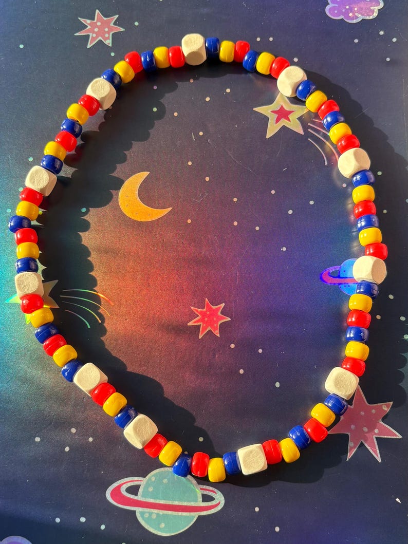 May include: A colorful beaded necklace with red, yellow, blue, and white beads. The necklace is arranged in a circular shape, with a space-themed background featuring stars, planets, and a crescent moon.