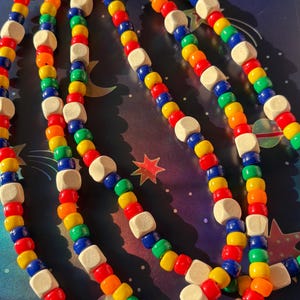 May include: A colorful beaded necklace with a rainbow of hues, including red, orange, yellow, green, and blue. The necklace features small, round beads interspersed with larger, square wooden beads. The beads are strung together, creating a playful and vibrant accessory.