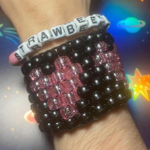 Puede incluir: Dos pulseras de cuentas. La pulsera superior tiene cuentas de letras blancas que deletrean "STRAWBEE". La pulsera inferior es negra con un diseño de corazón rosa. Las pulseras se usan en la muñeca.