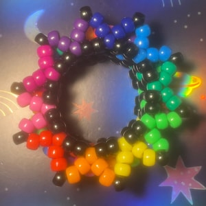 Pode incluir: Uma pulseira de contas coloridas com um design em forma de estrela. A pulseira apresenta um arco-&iacute;ris de cores, incluindo rosa, vermelho, laranja, amarelo, verde, azul e roxo, com contas pretas entre cada cor. A pulseira tem formato circular.