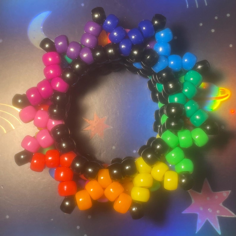 Pode incluir: Uma pulseira de contas coloridas com um design em forma de estrela. A pulseira apresenta um arco-&iacute;ris de cores, incluindo rosa, vermelho, laranja, amarelo, verde, azul e roxo, com contas pretas entre cada cor. A pulseira tem formato circular.
