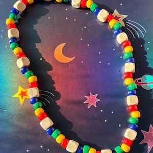May include: A colorful beaded necklace featuring a mix of small, round beads in red, yellow, green, and blue, interspersed with square, natural wood beads. The necklace is displayed on a space-themed background with stars and planets.