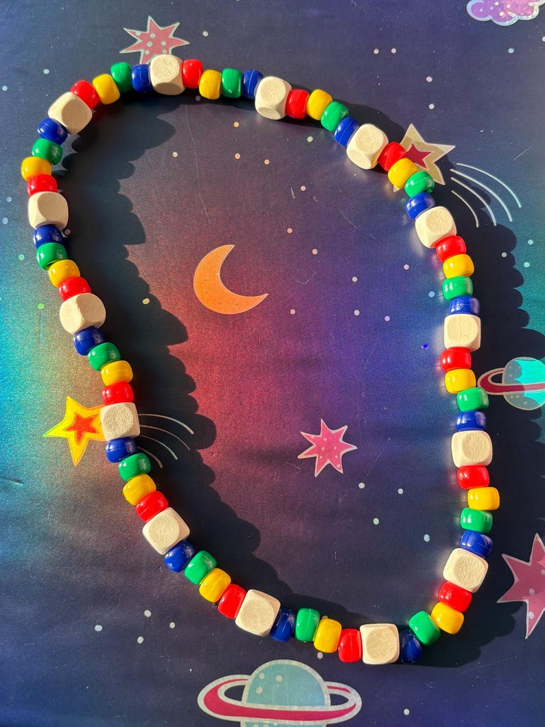 May include: A colorful beaded necklace featuring a mix of small, round beads in red, yellow, green, and blue, interspersed with square, natural wood beads. The necklace is displayed on a space-themed background with stars and planets.
