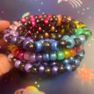 Pode incluir: Pilhas de pulseiras de contas coloridas. As pulseiras apresentam uma variedade de cores, incluindo roxo, azul, verde, amarelo, laranja, rosa e preto. As contas s&atilde;o enfiadas para criar um efeito em camadas. Um acess&oacute;rio divertido e vibrante.