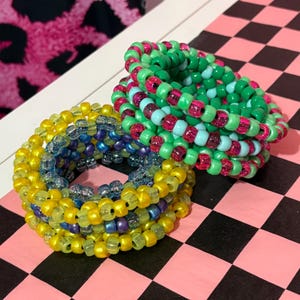 Pode incluir: Tr&ecirc;s pulseiras de contas em cores vibrantes. Uma pulseira &eacute; amarela e azul, outra &eacute; verde, rosa e azul, e a terceira &eacute; amarela e azul. As pulseiras est&atilde;o empilhadas sobre uma superf&iacute;cie quadriculada rosa e preta.
