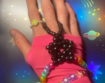 May include: A hand adorned with beaded jewelry. A smiley face ring, a black star-shaped bracelet, and a rainbow-colored bracelet with flower-shaped beads spelling "rawb". The hand is wearing a pink sleeve.