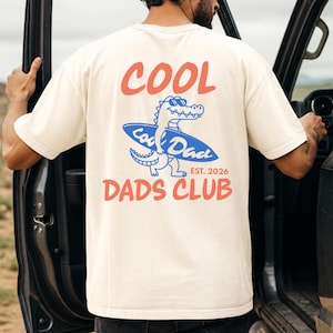 May include: Cream-colored t-shirt with a graphic on the back. The graphic features the words "COOL DADS CLUB" in red, with a cartoon alligator wearing sunglasses and riding a blue surfboard. The text "EST. 2026" is also included.