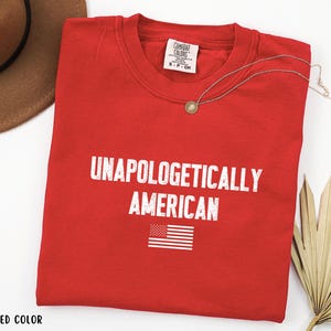 Może przedstawiać: Czerwony t-shirt z okrągłym dekoltem z białym napisem "UNAPOLOGETICALLY AMERICAN" i małą grafiką amerykańskiej flagi. Widoczny jest r&oacute;wnież złoty naszyjnik z wisiorkiem i brązowy kapelusz.