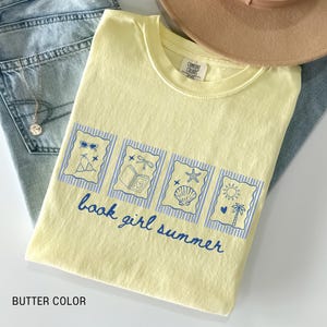 May include: A butter-colored t-shirt with a blue graphic that reads "book girl summer." The graphic features four framed images with summer-themed illustrations. The shirt is folded on top of a pair of light-wash jeans and a tan hat.