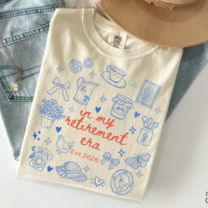 K&ouml;nnte beinhalten: Elfenbeinfarbenes T-Shirt mit blauem und rotem Grafikdesign. Das Design umfasst verschiedene Illustrationen wie Blumen, eine Teekanne, eine Tasse, ein Glas Marmelade und den Text "in my retirement era Est.2026."