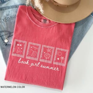 May include: A watermelon-colored t-shirt with a white graphic design featuring sunglasses, a book, a seashell, and a sun. The text "book girl summer" is written in white script. The shirt is laid on top of blue jeans and a tan hat.