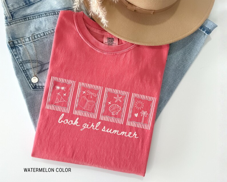 May include: A watermelon-colored t-shirt with a white graphic design featuring sunglasses, a book, a seashell, and a sun. The text "book girl summer" is written in white script. The shirt is laid on top of blue jeans and a tan hat.