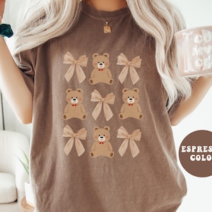 May include: A brown t-shirt with a graphic print of multiple brown teddy bears wearing red bows. The text "ESPRESSO COLOR" is printed in white on a brown circle in the bottom right corner of the image.