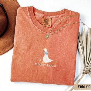May include: A coral coloured t-shirt with a white goose illustration and the text "Mother Goose".