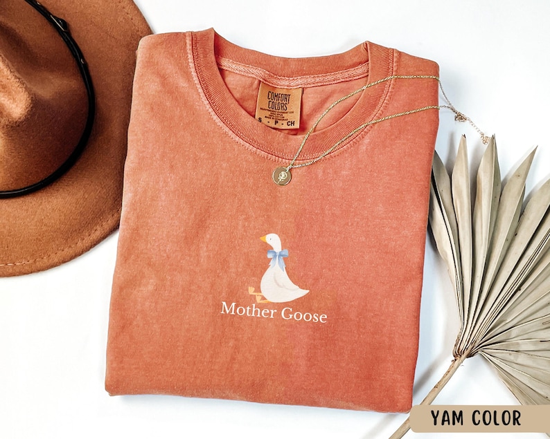 May include: A coral coloured t-shirt with a white goose illustration and the text "Mother Goose".