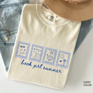 May include: Ivory-colored t-shirt with a blue graphic that says "book girl summer." The graphic includes images of sunglasses, a book, a seashell, and a sun. The shirt is laid on top of blue jeans and a tan hat.
