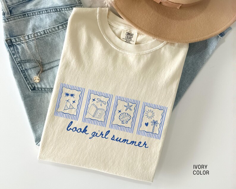 May include: Ivory-colored t-shirt with a blue graphic that says "book girl summer." The graphic includes images of sunglasses, a book, a seashell, and a sun. The shirt is laid on top of blue jeans and a tan hat.