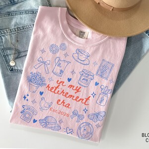 K&ouml;nnte beinhalten: Ein hellrosa T-Shirt mit blauen Illustrationen und dem Text "in my retirement era Est. 2026". Das T-Shirt liegt auf einer blauen Jeans und einem beigen Hut. Das Shirt zeigt Zeichnungen von Blumen, einer Teekanne und einem Schmetterling.
