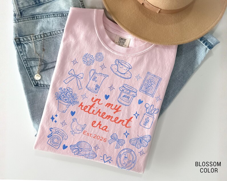 K&ouml;nnte beinhalten: Ein hellrosa T-Shirt mit blauen Illustrationen und dem Text "in my retirement era Est. 2026". Das T-Shirt liegt auf einer blauen Jeans und einem beigen Hut. Das Shirt zeigt Zeichnungen von Blumen, einer Teekanne und einem Schmetterling.