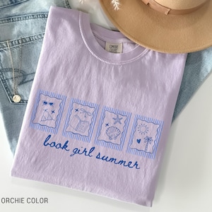 May include: A light purple t-shirt with a blue graphic that says "book girl summer." The graphic includes images of sunglasses, a beach chair, a book, a seashell, and a sun with a palm tree. The shirt is folded on top of blue jeans.