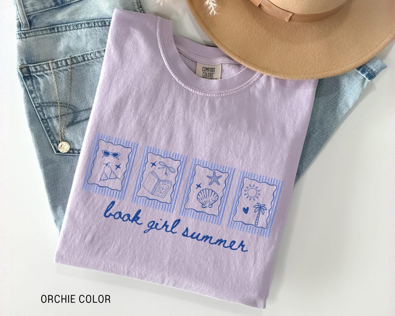 May include: A light purple t-shirt with a blue graphic that says "book girl summer." The graphic includes images of sunglasses, a beach chair, a book, a seashell, and a sun with a palm tree. The shirt is folded on top of blue jeans.