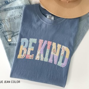 May include: A blue jean-colored t-shirt with the words "BE KIND" in a patchwork design. The shirt is folded on top of a pair of blue jeans and a tan hat. A gold necklace is visible on the jeans.