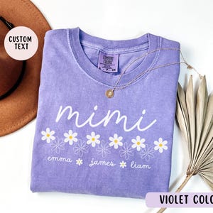 May include: Purple t-shirt with the word "mimi" and a row of white daisies. Below the daisies are the names "emma", "james", and "liam".