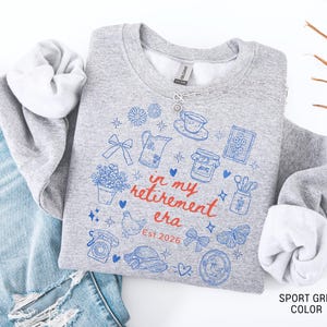 K&ouml;nnte beinhalten: Sportgraues Sweatshirt mit blauen und roten grafischen Illustrationen und dem Text "in my retirement era Est. 2026". Das Design umfasst eine Teekanne, Blumen und einen Schmetterling. Das Sweatshirt wird mit einer blauen Jeans gezeigt.