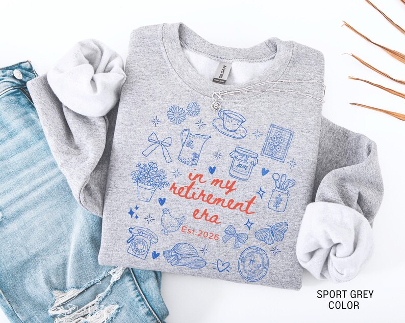 K&ouml;nnte beinhalten: Sportgraues Sweatshirt mit blauen und roten grafischen Illustrationen und dem Text "in my retirement era Est. 2026". Das Design umfasst eine Teekanne, Blumen und einen Schmetterling. Das Sweatshirt wird mit einer blauen Jeans gezeigt.