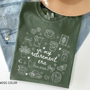 K&ouml;nnte beinhalten: Ein moosgr&uuml;nes T-Shirt mit einem wei&szlig;en Grafikdesign, das verschiedene Illustrationen zum Thema Ruhestand zeigt, darunter eine Teetasse, Blumen und ein Schmetterling. Der Text "in my retirement era" und "Est. 2026" sind ebenfalls enthalten. Das Shirt liegt auf einer Jeans.