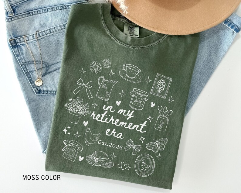 K&ouml;nnte beinhalten: Ein moosgr&uuml;nes T-Shirt mit einem wei&szlig;en Grafikdesign, das verschiedene Illustrationen zum Thema Ruhestand zeigt, darunter eine Teetasse, Blumen und ein Schmetterling. Der Text "in my retirement era" und "Est. 2026" sind ebenfalls enthalten. Das Shirt liegt auf einer Jeans.