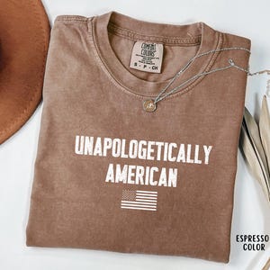 Może przedstawiać: Koszulka w kolorze espresso z białym napisem "UNAPOLOGETICALLY AMERICAN" i małą grafiką amerykańskiej flagi. Koszulka ma okrągły dekolt i kr&oacute;tkie rękawy. Srebrny naszyjnik z wisiorkiem leży na koszulce.