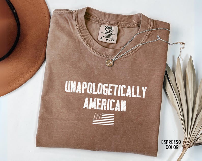 Może przedstawiać: Koszulka w kolorze espresso z białym napisem "UNAPOLOGETICALLY AMERICAN" i małą grafiką amerykańskiej flagi. Koszulka ma okrągły dekolt i kr&oacute;tkie rękawy. Srebrny naszyjnik z wisiorkiem leży na koszulce.