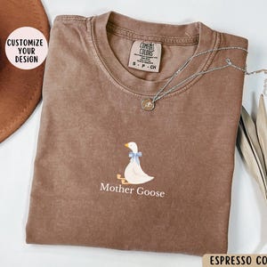 May include: A brown t-shirt featuring a white goose illustration and the text "Mother Goose". The shirt is made from a soft, comfortable material and has a distressed look. The text "COMFORT COLORS" is visible on the tag.
