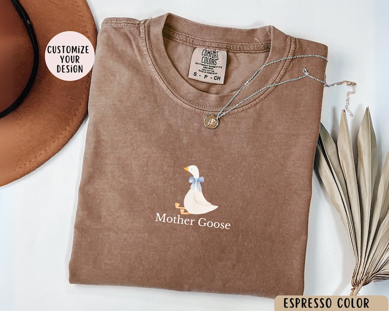 May include: A brown t-shirt featuring a white goose illustration and the text "Mother Goose". The shirt is made from a soft, comfortable material and has a distressed look. The text "COMFORT COLORS" is visible on the tag.