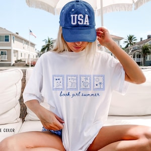 May include: White t-shirt with a blue graphic of four summer-themed images and the text "book girl summer." The shirt is paired with a blue baseball cap with "USA" in white letters.