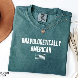 Może przedstawiać: Niebieska koszulka Comfort Colors z napisem "UNAPOLOGETICALLY AMERICAN" w białych literach i małą amerykańską flagą. Złoty naszyjnik i brązowy kapelusz r&oacute;wnież znajdują się na zdjęciu.