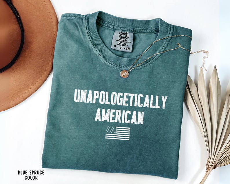Może przedstawiać: Niebieska koszulka Comfort Colors z napisem "UNAPOLOGETICALLY AMERICAN" w białych literach i małą amerykańską flagą. Złoty naszyjnik i brązowy kapelusz r&oacute;wnież znajdują się na zdjęciu.