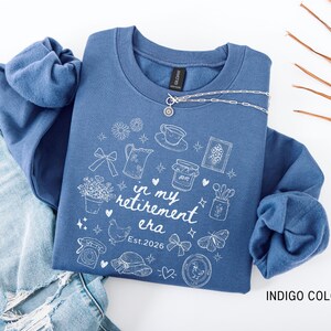 K&ouml;nnte beinhalten: Indigoblaues Sweatshirt mit wei&szlig;en grafischen Illustrationen und dem Text "in my retirement era Est. 2026". Eine silberne Halskette liegt &uuml;ber dem Sweatshirt. Das Sweatshirt wird mit einer blauen Jeans kombiniert.