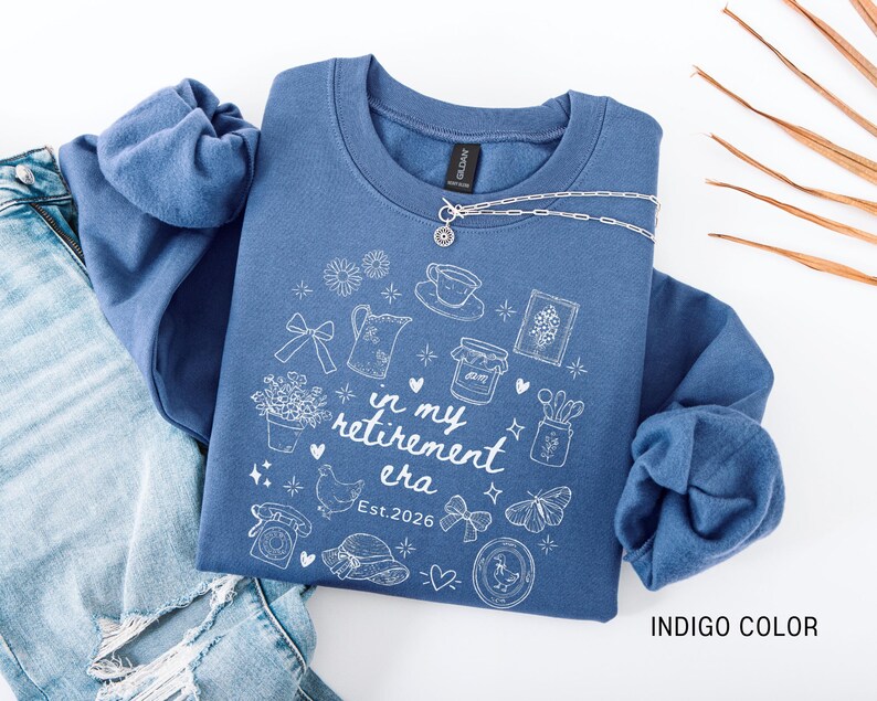 K&ouml;nnte beinhalten: Indigoblaues Sweatshirt mit wei&szlig;en grafischen Illustrationen und dem Text "in my retirement era Est. 2026". Eine silberne Halskette liegt &uuml;ber dem Sweatshirt. Das Sweatshirt wird mit einer blauen Jeans kombiniert.