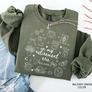 K&ouml;nnte beinhalten: Milit&auml;rgr&uuml;nes Sweatshirt mit wei&szlig;en grafischen Illustrationen und dem Text "in my retirement era Est.2026". Das Design umfasst eine Teekanne, Blumen, ein Huhn und andere skurrile Elemente. Eine silberne Halskette liegt auf dem Sweatshirt.