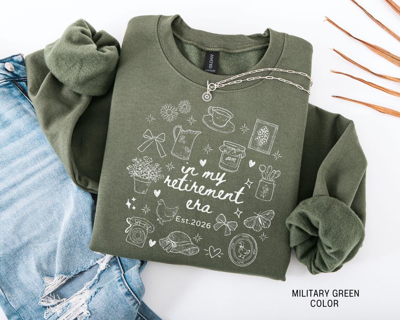 K&ouml;nnte beinhalten: Milit&auml;rgr&uuml;nes Sweatshirt mit wei&szlig;en grafischen Illustrationen und dem Text "in my retirement era Est.2026". Das Design umfasst eine Teekanne, Blumen, ein Huhn und andere skurrile Elemente. Eine silberne Halskette liegt auf dem Sweatshirt.
