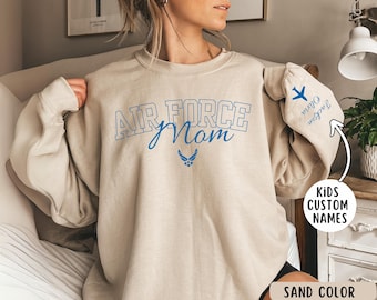 May include: Sand colored sweatshirt with the text "Air Force Mom" in blue lettering. The sweatshirt has a blue Air Force logo and a space for a child's name on the sleeve.